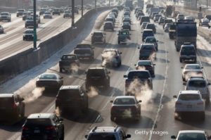 Four lanes of traffic, with several cars emitting visible exhaust gases.