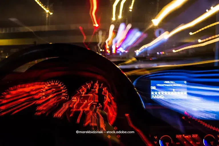 The driver's vision is impaired, as shown through the blurry visuals of the dashboard, radio display and field of vision beyond the windshield. Nighttime streetlights are blurred.