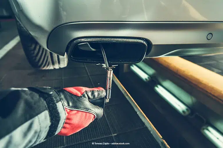 A mechanic with red gloves uses a tool to inspect the tailpipe of a vehicle.