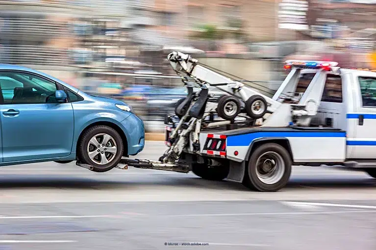 A blue and white tow truck tows a blue car. The blurry background indicates that the tow truck is speeding down the street.