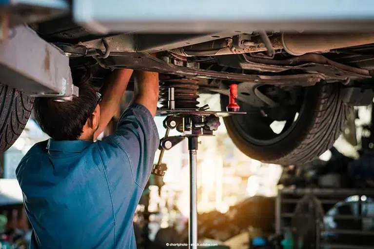 A mechanic stands underneath a car suspended by a car lift. He manually adjusts components currently obscured from view.