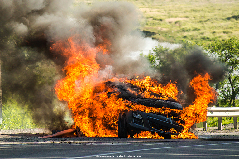 A vehicle on the side of a highway is engulfed in flames, emitting black and white smoke.