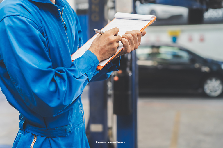 A mechanic holds an orange clipboard. He uses a pen to cross off items on a checklist.