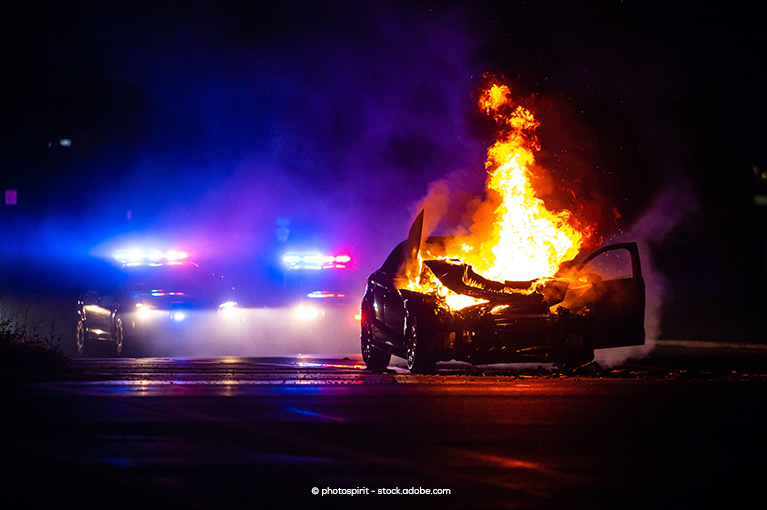 It is nighttime. A single vehicle has flames erupt from its engine compartment. Behind the flaming vehicle are two police cars with police lights alight.