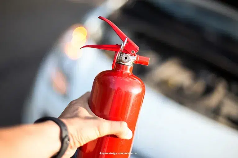 A person wearing a watch is holding a fire extinguisher. In the blurred background is a car with the hood open.