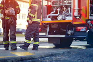 Two firefighters stand beside a fire truck, parked on the side of the road in the daytime.