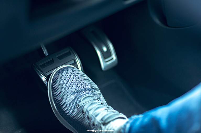 A close-up shot of a depressed brake pedal. A driver wearing sneakers presses down on the brake pedal.