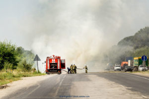 White smoke indicates a fire on the street, close to it. A firetruck is parked on the left side of the road. Three firefighters stand beside it, with one holding a fire hose. Vehicles parked in the opposite direction populate the right side of the road.