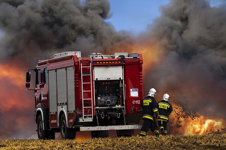 Black smoke fills the background of the image. In the foreground, a red firetruck is parked. Two firefighters stand to its right, attempting to put out a nearby fire.