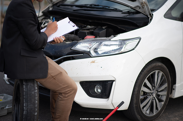 A man with a black blazer and brown pants checks items of a list clipped to a clipboard. He sits on an upright tire in front of a white vehicle with an open hood.