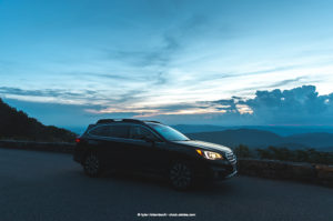 It is sunset, with clouds obscuring the sun. In the dimly lit view, a Subaru SUV is parked on the side of the road, wherein the road is demarcated by a cobblestone boundary.