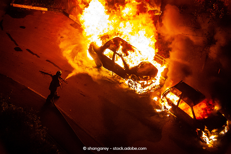 A single firefighter uses a hose to put out two car fires. The vehicle at the bottom corner of the image has flames throughout its cabin. However, the second vehicle in the middle of the image is completely engulfed in flames.