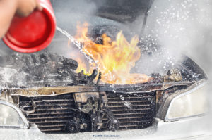 A person sprinkles water on a fire coming from a vehicle engine compartment.