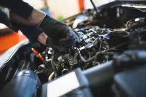 A mechanic with gloved hands manipulates a few small components in the engine compartment under the hood.