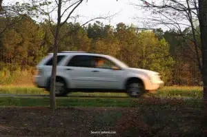 A white Nissan SUV blurs while driving on a paved road surrounded by patchy grass and forested areas.