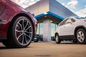 Two vehicles are parked in front of a dealership. A white SUV occupies the right side of the image. On the left side, only the red vehicle's passenger side front wheel is visible.