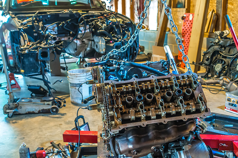 An engine is suspended by blue metal chains inside an auto shop. The vehicle that once housed the engine is being taken apart.