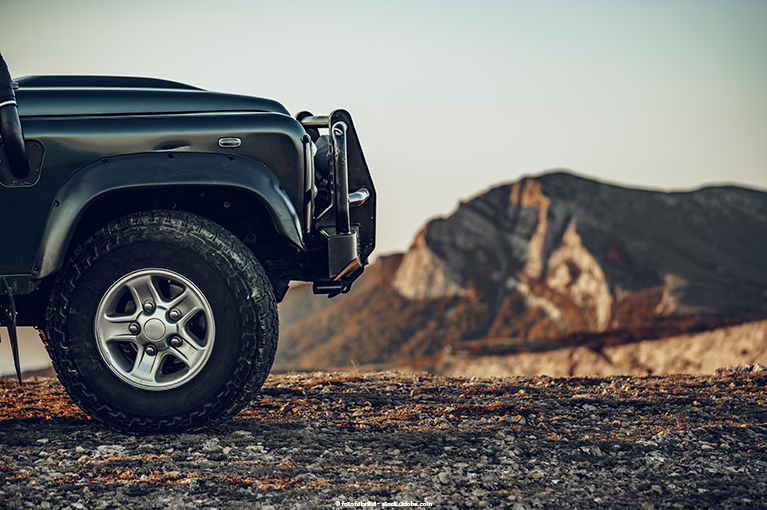 The front of a Jeep takes up the left half of the image. The Jeep itself is parked on a sandy, rocky top of a mountain.