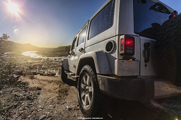 A grey Jeep Wrangler is parked off road in the muddy shore of a river.