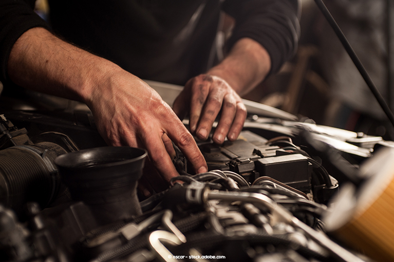 A bare-handed mechanic touches various components under the hood of a car.