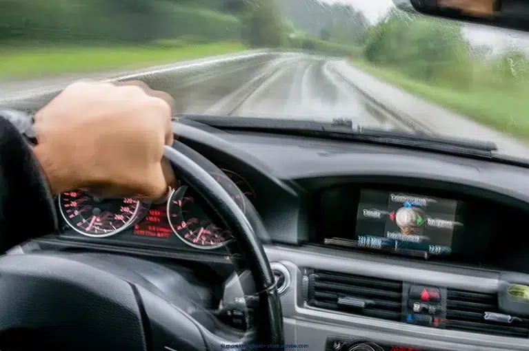 An image of the steering wheel, dashboard and single-lane highway from the perspective of the driver. The driver holds the steering wheel with his left hand. The image appears to be wobbly, as if the driver is cross-eyed or has blurry vision.