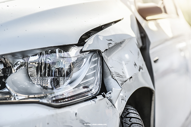 A close up of a headlight of a white vehicle. The body surrounding the headlight appears to be scraped and crumpled, indicating a collision.