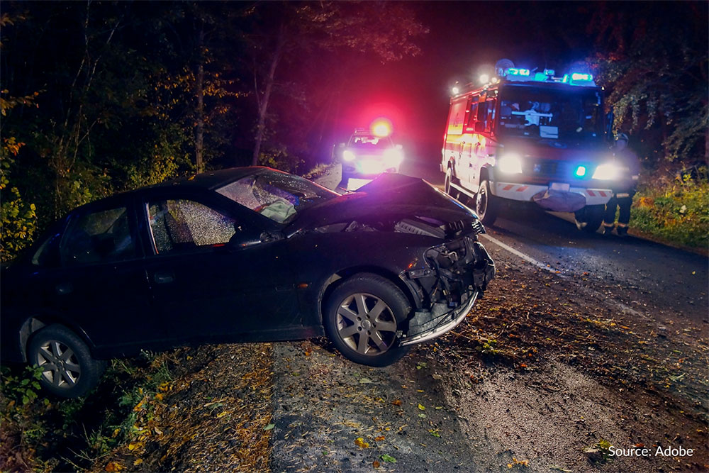 A black sedan has its windows shattered and its front hood and bumper destroyed. The sedan has rolled off the side of the road. Two emergency vehicles are parked on the road.