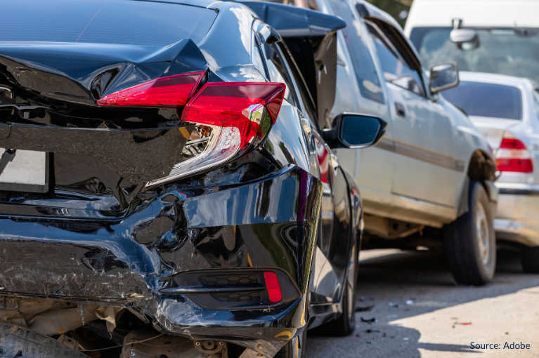 A pile-up crash has occurred. In the foreground of the image, a black car appears to have been rear-ended. It has also had its front hood crumpled. In between the black car and a grey car is a blue truck appearing to have been crushed.