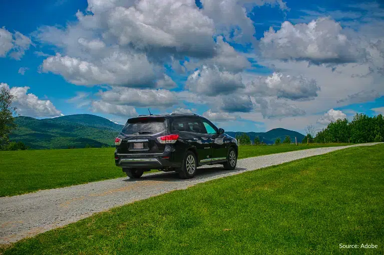 A black SUV is parked on a one-lane dirt road in the middle of a grassy field. It is daytime and clouds litter the bright blue sky.