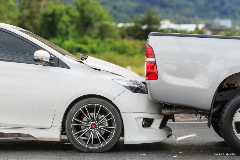 A small white car rear ended a white/grey pickup truck. Its frontal hood is broken, its headlights are broken, and its bumper is wedged underneath the truck's trunk.