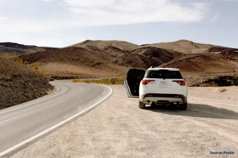 A white GMC vehicle is parked on a dirt turnabout of a highway in the desert. The GMC's driver side front door is open.