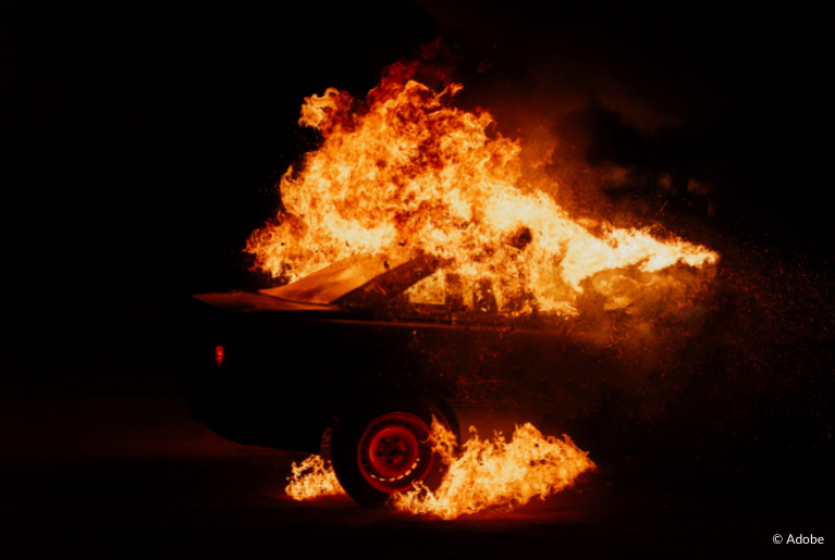 A car is engulfed in flames. The fire itself does not illuminate its surroundings. It is presumably nighttime.