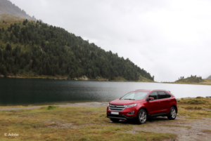 A red Ford Explorer SUV is parked on the shore of a lake. On the opposite side of the lake are forested hillsides.