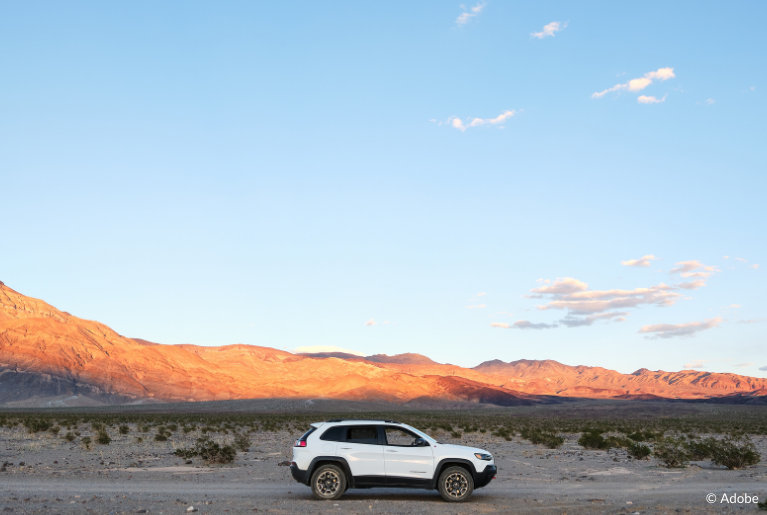 A white Jeep SUV is parked on an unpaved road in the middle of a desert, in the foreground of the image. In the background are illuminated mountains surrounding a shrub-laden plain.