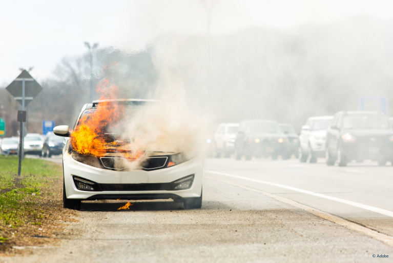 A fire erupts in the engine compartment of a white car parked on the side of a highway.