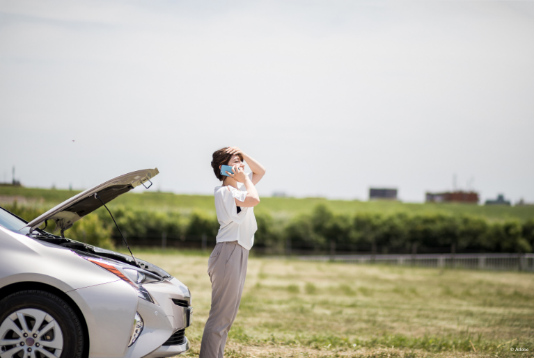 A woman stands in front of her car, holding her head in frustration as she talks on the phone. The silver car behind her has its hood popped open.