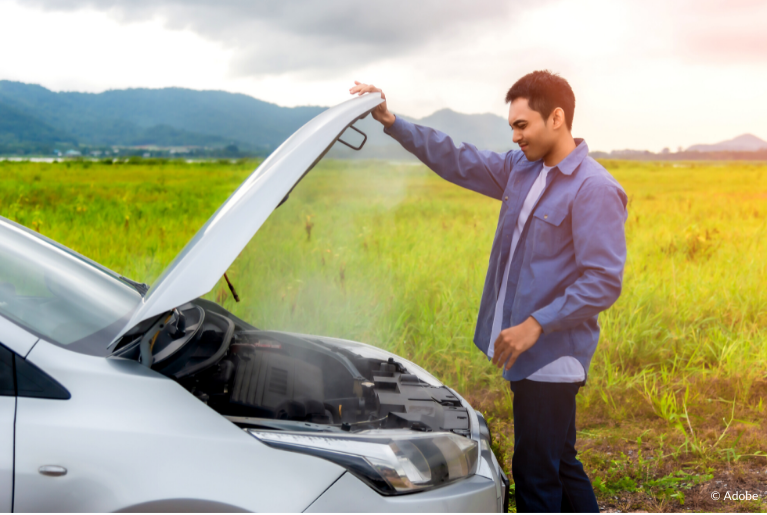 A man holds open the hood of his car. Light smoke is coming out of the engine compartment. Behind him and the car is a grassy field.