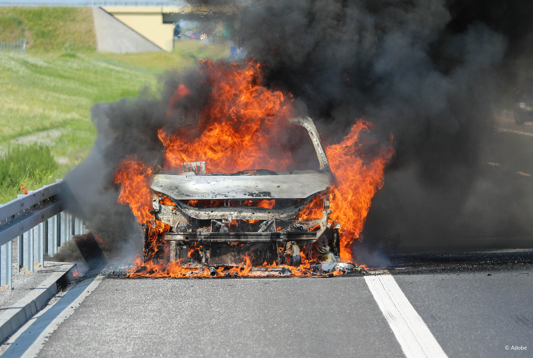 A vehicle is completely burned through, its metal parts exposed. A fire still engulfs the car. It is on the side of the road, next to highway guardrails.