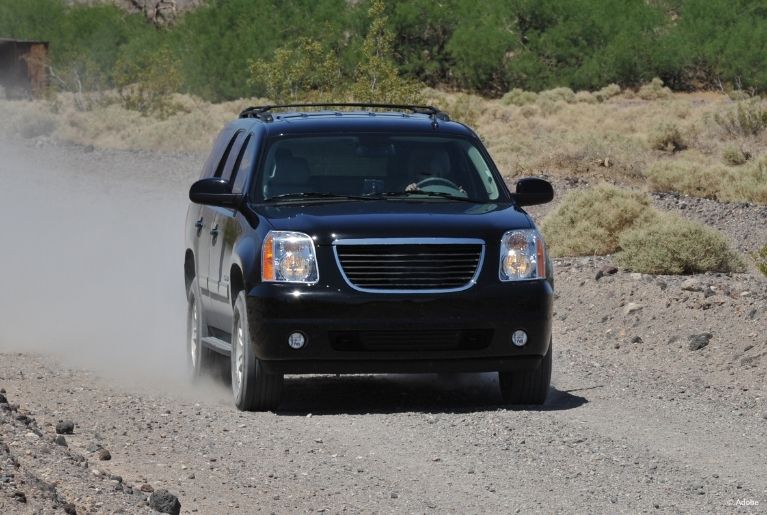 A black GMC Yukon kicks up dust behind it as it drives slowly on a dirt road.