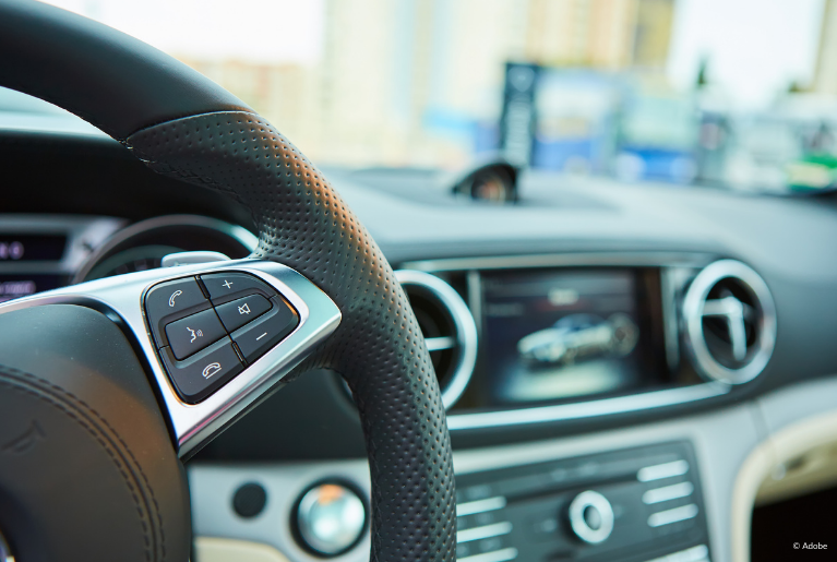 A close-up of buttons on a steering wheel. Out of focus, a radio display shows an image of a sedan.