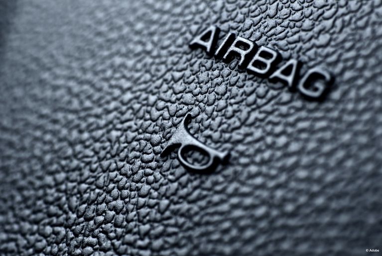 A close-up of the surface of a car horn, with indented text that reads "AIRBAG." An indent logo of a trumpet - representing the car horn noise - is positioned below the text.