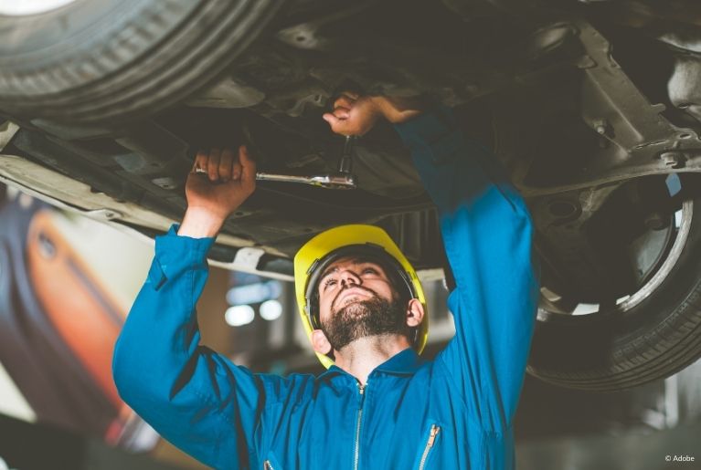 A mechanic in a blue jumpsuit and a yellow hardhat uses a torque wrench to unscrew a bolt under the car, which is suspended on a car lift.