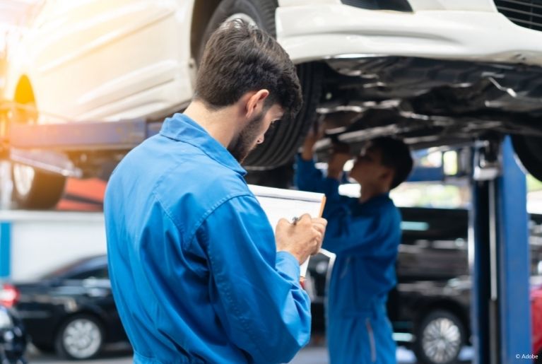 A mechanic in a blue jumpsuit crosses off items on a list clipped to a brown clipboard. A white vehicle is suspended on a car lift, while another mechanic inspects the suspension.