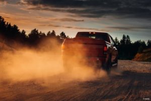 A red pickup truck kicks up dust behind it while driving on a dirt road at sunset.