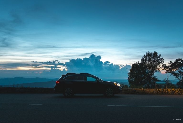 A Subaru SUV is parked on the side of a road at dusk. The sunset is obscured by low-level clouds. The SUV's headlights only partly illuminate the rock guardrail of the road.