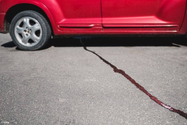 A thin stream of fluid leaks from a red vehicle parked in an outdoor lot.