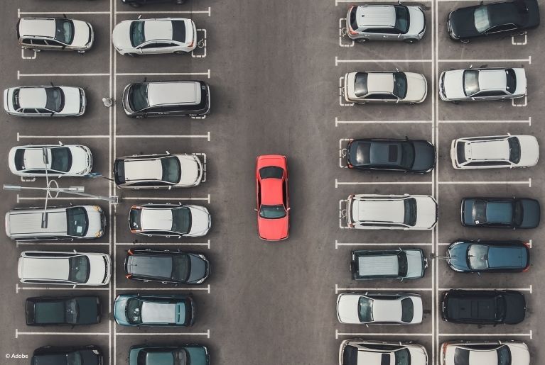 A red car drives down a parking lot filled with black, blue and grey vehicles.