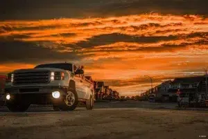 A white GMC truck parked on an empty paved road. The sunset illuminates the overcast sky in orange light. Behind the car are houses lining up the paved road on either side.