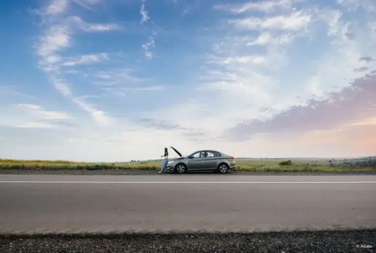 A woman is standing in front of her parked grey sedan, which has its hood up. Nissan Maxima cars reportedly experience issues with its CVT transmission.