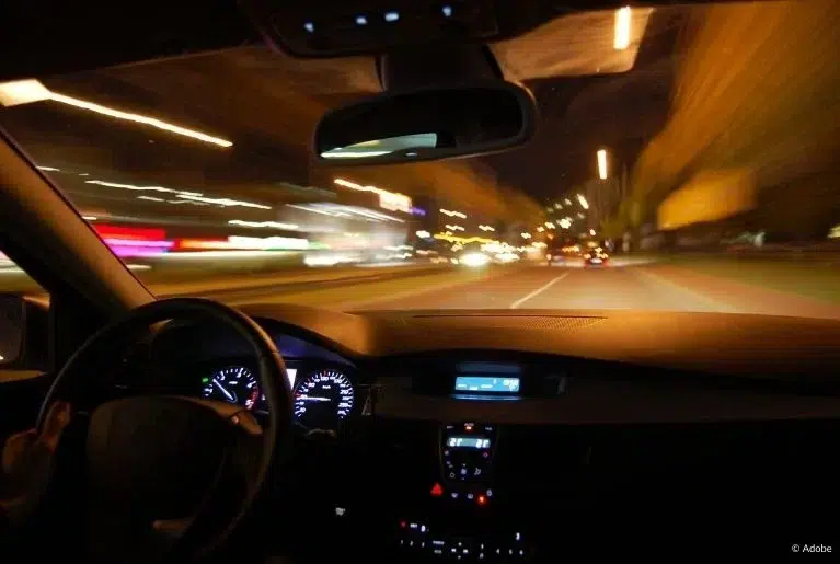 A front passenger seat shot of a steering wheel, dashboard, radio display and view of the street. Streetlights and other lights blur as the vehicle speeds through.